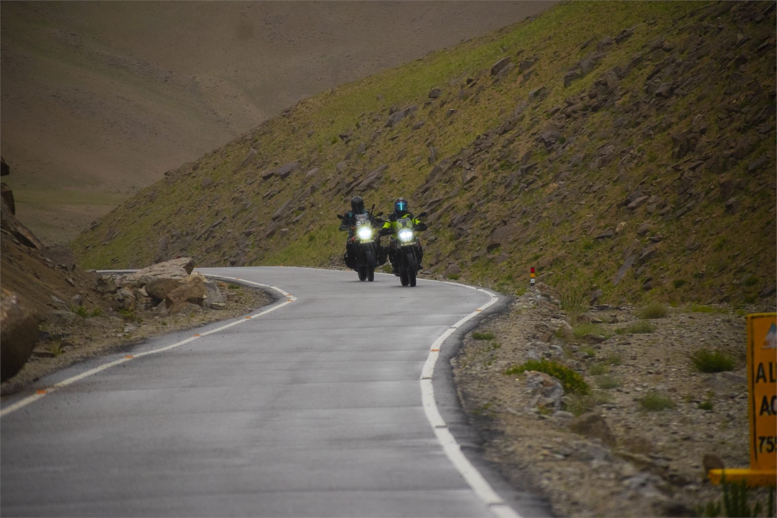 Two riders on Royal Enfields climbing a paved mountain road in Ladakh below a bare scree slope